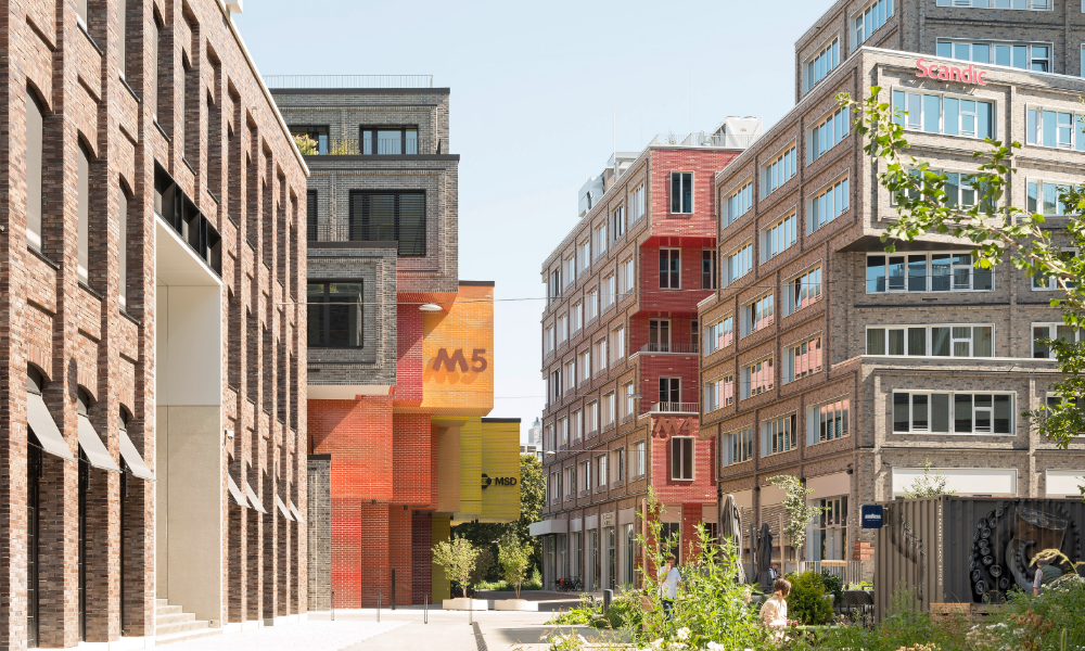 Moderner Hof zwischen Gebäuden mit markanten Fassaden, Gehweg mit Passanten und Sitzbereich im Freien.
