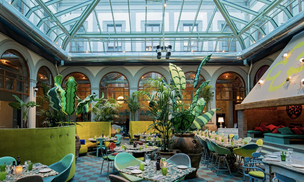 Innenansicht eines Restaurants in einem Wintergarten mit Glasdach, bunten Stühlen, gedeckten Tischen und großen Pflanzen.
