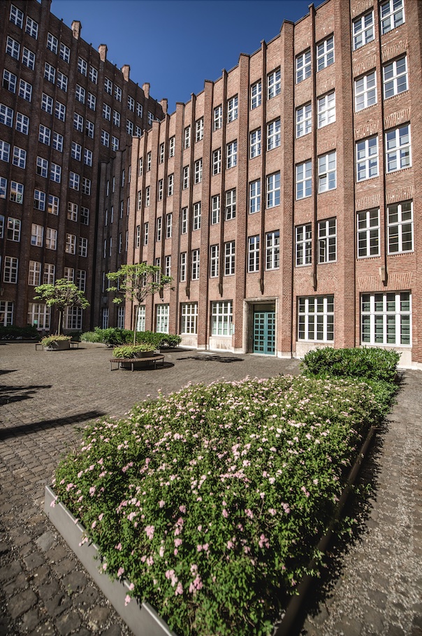 Innenhof mit roten Backsteinfassaden, vielen Fenstern, Blumenbeeten und kleinen Bäumen.